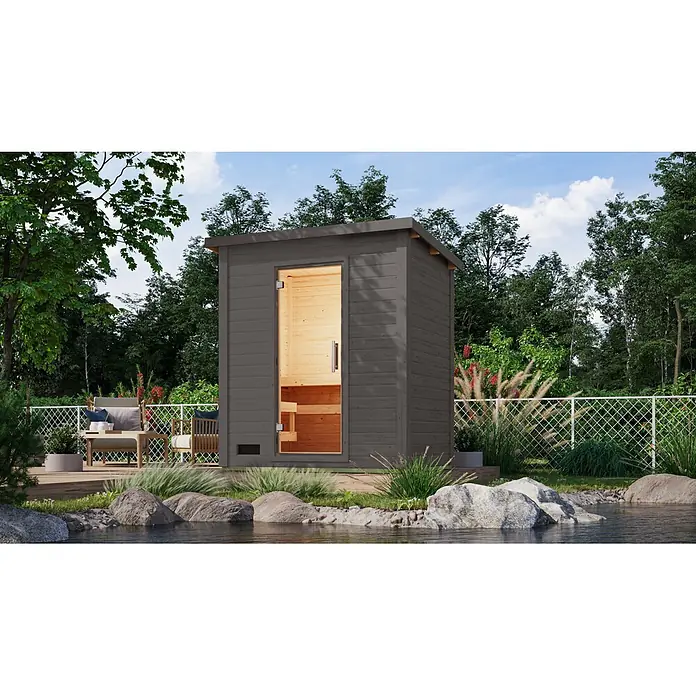 Karibu Saunahaus Kaapi 2 - 9 KW Ofen mit interner Steuerung, Anthrazit, L x B x H: 146 x 196 x 228 cm - Dunkelgraue Holz-Außensauna mit Glastür und hellem Interieur, am Wasser in einem Garten.