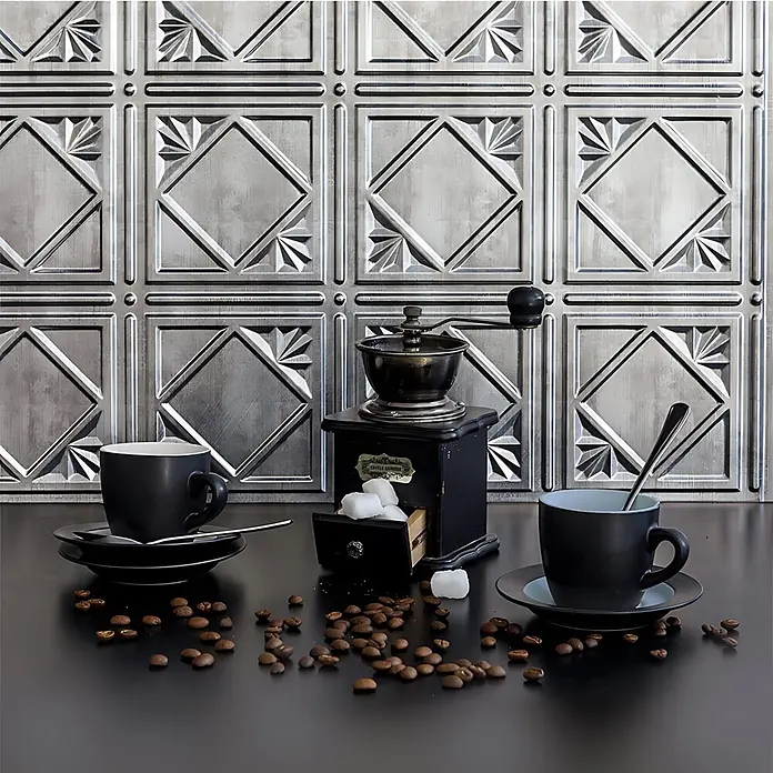 Profhome Wandpaneel 3D 705632 Artnuovo Crosshatch Silver Dekorpaneel Vintage silber 1,7 m2 - 470 x 610 mm - Zwei schwarze matte Kaffeetassen mit Untertassen und eine vintage Kaffeemühle auf einer dunklen Arbeitsplatte, umgeben von Kaffeebohnen und Zuckerwürfeln.