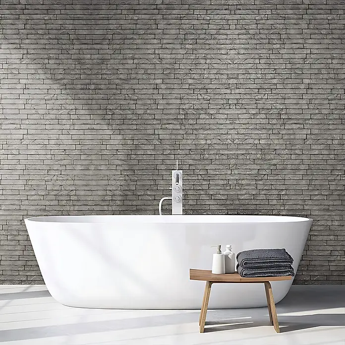 Profhome Wandpaneel 3D 704500 Ledge Stone Portland Cement Dekorpaneel Stein Optik grau 2 m2 - 610 x 600 mm - Weiße freistehende Badewanne in einem modernen Badezimmer. Daneben ein Holzhocker mit Handtüchern und Flaschen.