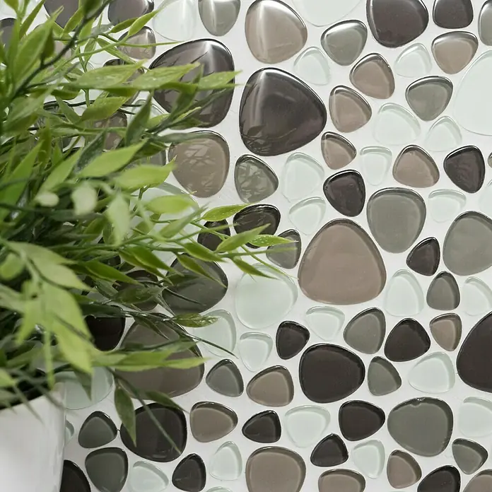 Mosaikfliese Kiesel Crystal Mix XCM PG66 - 29,9 x 29,9 cm, Braun/Beige/Weiß, Glänzend - Nahaufnahme eines Mosaiks aus polierten Kieselsteinen in Grau-, Braun- und Weißtönen, teilweise von einer Zimmerpflanze verdeckt.