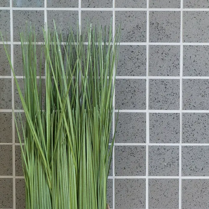 Mosaikfliese Quadrat Artifical XCM ASM43 - 30,5 x 30,5 cm, Grau, Glänzend - Nahaufnahme von grünem Ziergras vor einem Hintergrund aus grauen Fliesen, ideal für moderne Dekorationen.