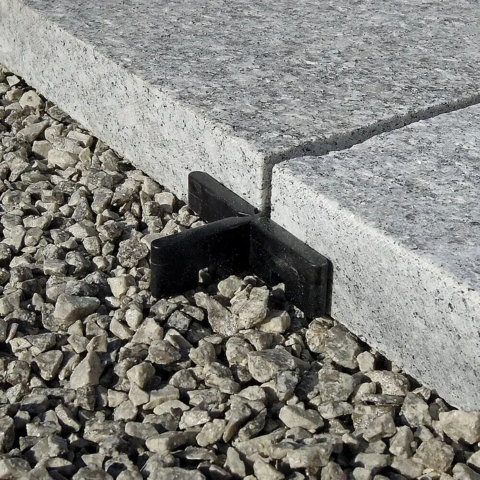 Fliesenkreuz FK - 50 Stk., Breite: 0,3 cm, Kunststoff - Das Bild zeigt einen schwarzen Fugenkreuz für Terrassenplatten, der zwischen grauen Steinplatten auf Kies platziert ist. Ideal für gleichmäßige Fugen.