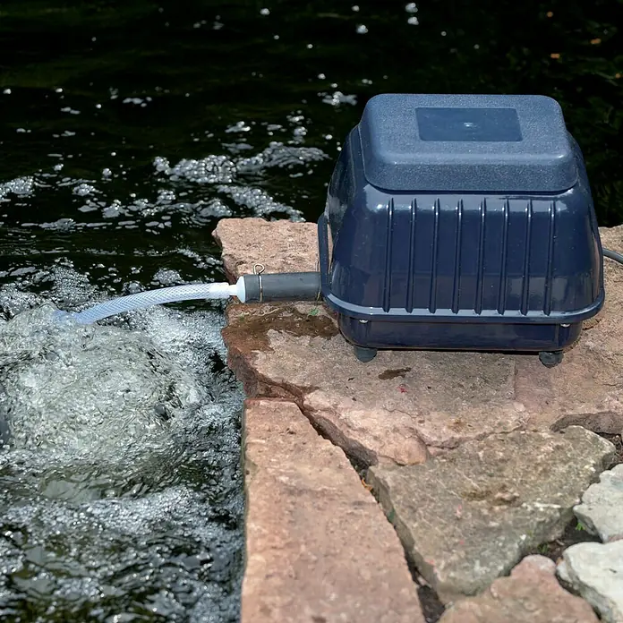Ubbink Teichbelüfter Air 4000 outdoor - 33 W, Teichgröße: 4 m³ - 15 m³ - Teichbelüfter auf einem Steinsockel am Ufer, sorgt für Sauerstoff im Wasser. Das Gerät ist kompakt und wetterfest konstruiert für den Außeneinsatz.