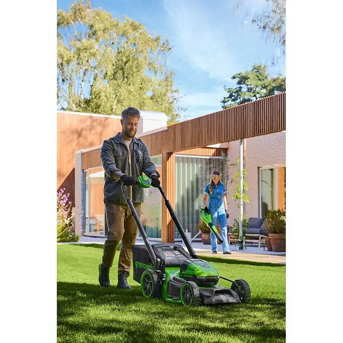 Greenworks Akku-Rasenmäher Gen2 GW 2X24V BL 46 cm - 48 V, 2 Akkus, 4 Ah, Schnittbreite: 33 mm - Ein Mann mäht den Rasen mit einem grün-schwarzen Greenworks Rasenmäher, während eine Frau einen Laubbläser in der Nähe benutzt.
