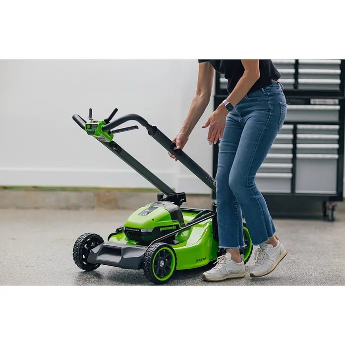 Greenworks Akku-Rasenmäher Gen2 GW 2X24V BL 46 cm - 48 V, 2 Akkus, 4 Ah, Schnittbreite: 33 mm - Eine Person stellt den Griff eines leuchtend grünen Greenworks Elektro-Rasenmähers in einer Garage ein.