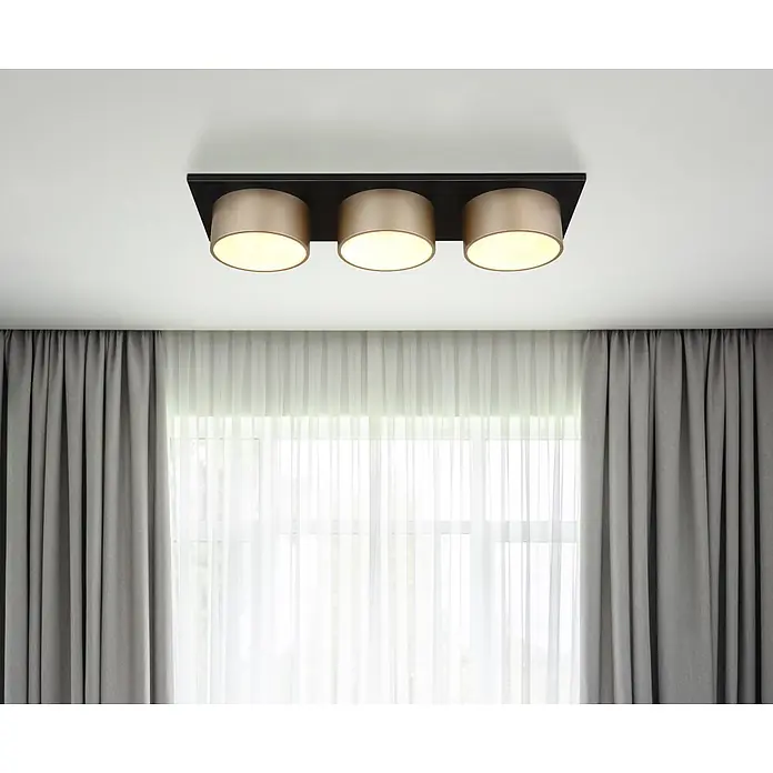 Globo Deckenleuchte Raymond - 24 W, L x B x H: 580 x 200 x 110 mm, Lichtfarbe: Warmweiß - Drei goldfarbene zylindrische Deckenstrahler auf schwarzer Rechteckbasis, über weißen Gardinen und grauen Vorhängen montiert.