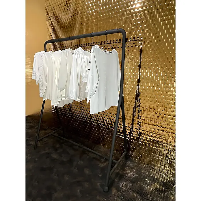 Wallface Wandpaneel 24955 RATTAN 20 Gold Wandverkleidung 3D glänzend gold 2,6 m2 - 2.600 x 1.000 mm - Weiße Blusen und Hemden hängen an einem schwarzen Kleiderständer vor einer schimmernden, goldenen Strukturwand.