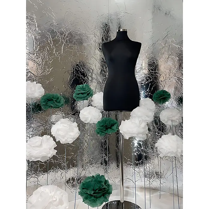 Wallface Wandpaneel 24945 CREPA Silver Wandverkleidung 3D spiegelnd silber 2,6 m2 - 2.600 x 1.000 mm - Schwarze Schaufensterpuppe auf Chromständer, umgeben von weißen und grünen Papierblumen vor silbern zerknittertem Hintergrund.