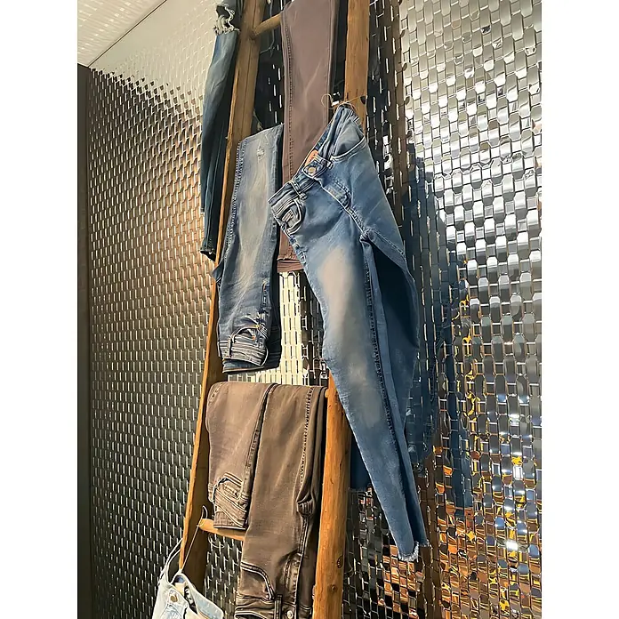 Wallface Wandpaneel 24956 RATTAN 20 Silver Wandverkleidung 3D glänzend silber 2,6 m2 - 2.600 x 1.000 mm - Sortiment blauer und grauer Jeans aus Denim, auf einer rustikalen Holzleiter vor einer strukturierten, reflektierenden Wand.
