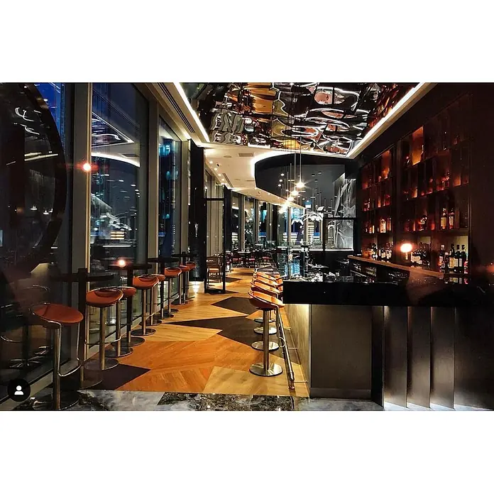 Wallface Wandpaneel 27049 OCEAN Anthracite Wandpaneel Metall-Optik spiegelnd grau 2,6 m2 - grau-braun - Eine elegante Bar mit langem Tresen, hohen Hockern und Flaschenregalen, vor großen Fenstern mit Blick auf eine nächtliche Stadt.