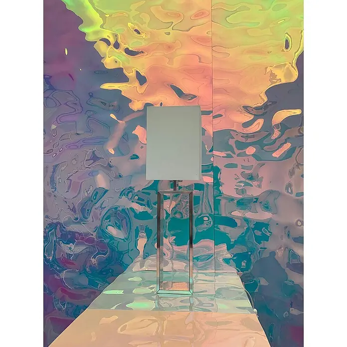 Wallface Wandpaneel 27822 OCEAN Hollywood Blue Wandpaneel Metall-Optik holographisch 2,6 m2 - blau-lila, 2.600 x 1.000 mm - Moderne Tischlampe mit weißem quadratischem Schirm und Chromfuß, auf einer reflektierenden Fläche vor irisierendem, welligem Hintergrund.