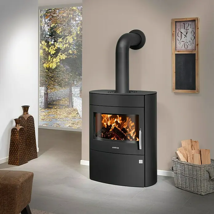 Justus Kaminofen Agero 2.0 - 7 kW, Raumheizvermögen: 144 m³, Material Verkleidung: Stahl, Schwarz - Ein moderner, schwarzer Kaminofen der Marke Justus mit brennendem Feuer im Inneren, der eine gemütliche Atmosphäre schafft. Holzkorb und Dekoration.