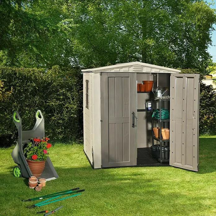 Keter Caseta de jardín Factor 6x6 - Medidas ext. (An x Pr): 195,5 x 178 cm, Taupe/Beige, Plástico, Área: 3,47 m² - Escena de jardín con caseta, carretilla, plantas y herramientas de jardín en el césped delante de árboles.
