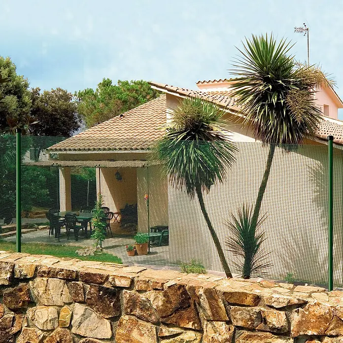 Nortene Malla protectora Cuadranet 11 - Verde, L x Al: 5 x 1 m - Una valla de malla metálica verde rodea una casa con un jardín de rocas en el patio delantero. La valla es una solución robusta y duradera.