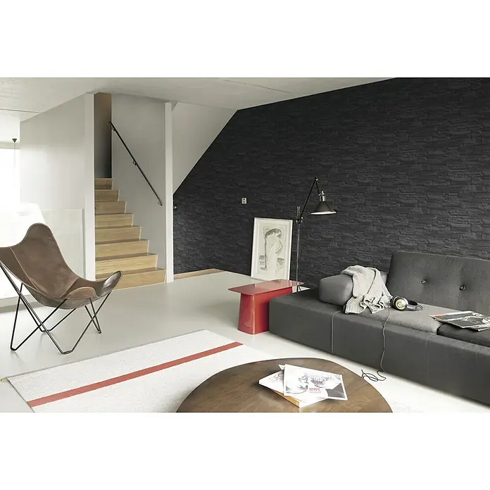 Rasch Vliestapete Steinoptik - Grau, 3D-Optik, 10,4 x 0,53 m - Moderne Wohnraumgestaltung mit dunkler Akzentwand und Ledersessel. Das Interieur besticht durch stilvolle Möbel und ein minimalistisches Design.