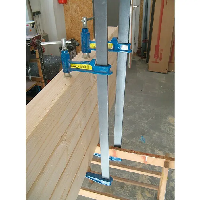 Piher Sargento F-40 - Anchura de sujeción: 400 mm, Fuerza de tensión: 9.000 N, Apertura máxima: 120 mm - Primer plano de tablones de madera asegurados con abrazaderas verticales azules sobre una estructura de madera, en un taller.