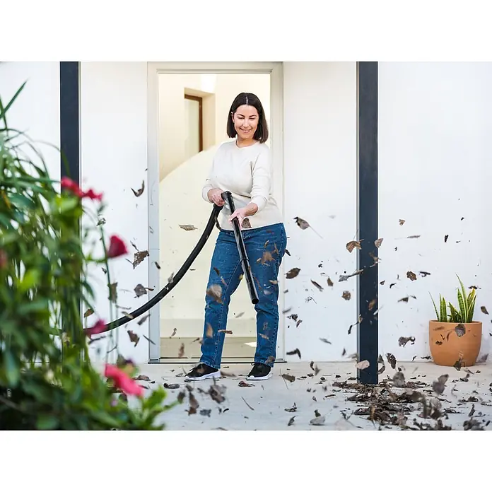 Nilfisk Aspirador en seco y húmedo eléctrico Buddy II 18 - 1.200 W, Volumen de recipiente: 18 l, Con bolsa - Una mujer usa un soplador/aspirador de hojas negro para limpiar hojas secas de un patio.