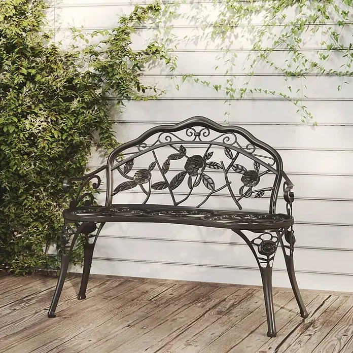 Furnicato Gartenbank Aluminiumguss - B x T x H: 100 x 54 x 80 cm, Schwarz - Dunkle Metall-Gartenbank mit Rosenmuster-Rückenlehne auf Holzterrasse, vor weißer Wand mit Kletterpflanzen.
