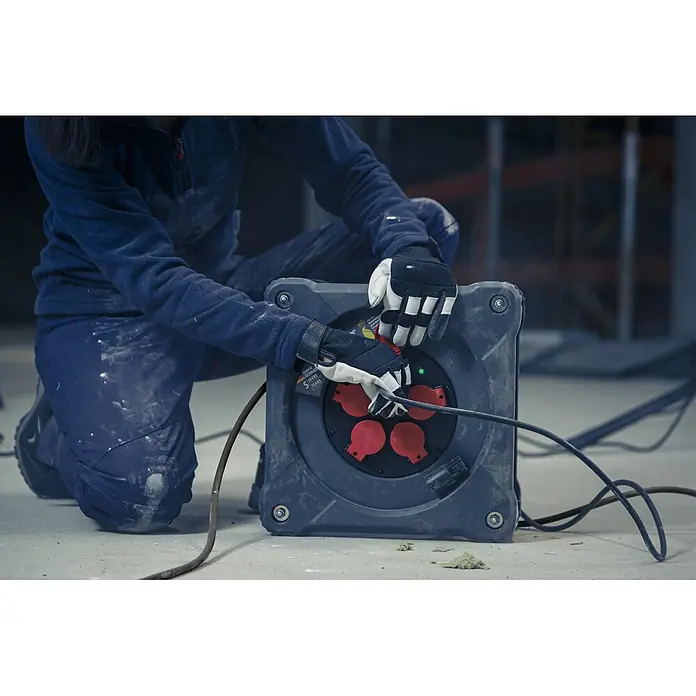 Brennenstuhl Kabeltrommel mit Powerblock KC 3200 5 - Kunststoff, 38 m, H07RN-F, IP44 - Ein Arbeiter mit Handschuhen verbindet ein Kabel mit einer robusten, dunkelgrauen Kabeltrommel mit roten Steckdosen auf einer Baustelle.