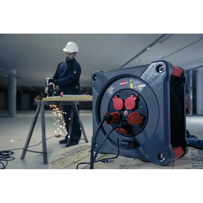 Brennenstuhl Kabeltrommel KA 3100 5 - Kunststoff, 33 m, H07RN-F, IP44 - Dunkelgraue Brennenstuhl Kabeltrommel mit roten Steckdosen, versorgt einen Arbeiter, der einen Winkelschleifer auf einer Baustelle benutzt.