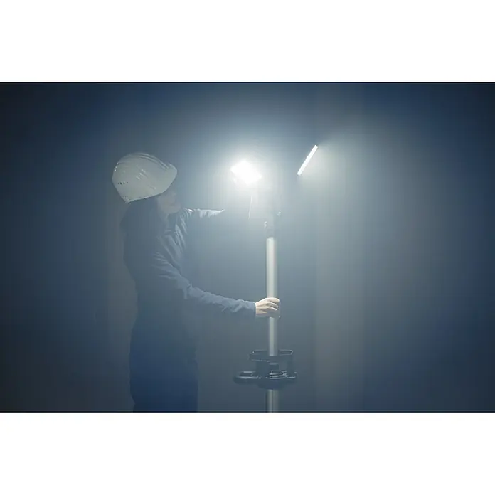 Brennenstuhl Mobiler LED-Strahler Dreikopf Stativstrahler - 100 W, L x B x H: 26 x 21 x 106 cm, IP34 - Person in Schutzhelm justiert Baustrahler auf Stativ, erhellt dunklen Raum hell.