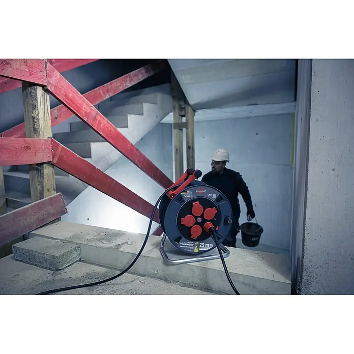 Brennenstuhl Kabeltrommel 300 KU 2200 - Kunststoff, 25 m, H07RN-F, IP44 - Eine brennenstuhl® 25m Kabeltrommel mit roten Steckdosen auf Beton auf einer Baustelle. Ein Arbeiter ist im Hintergrund.
