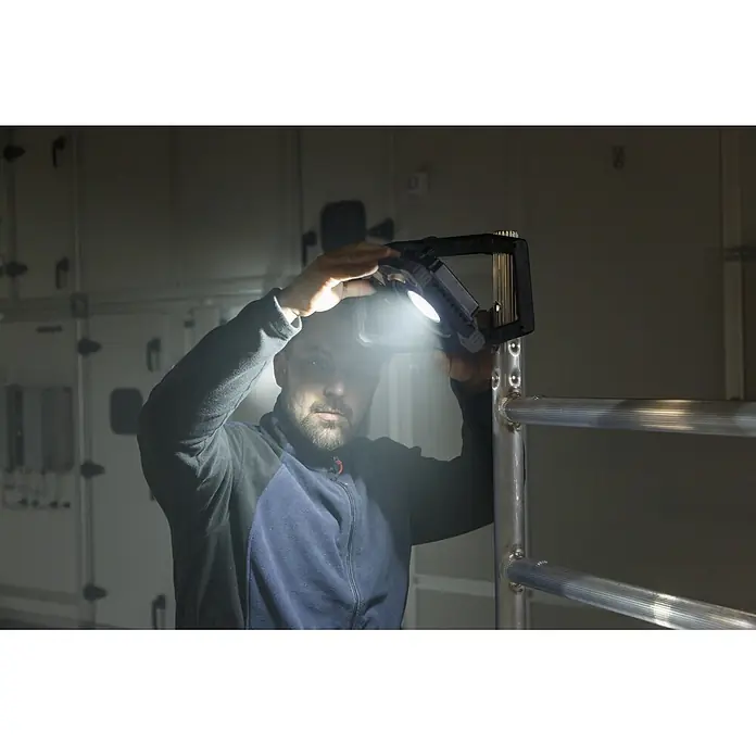 Brennenstuhl LED-Baustrahler - 6.800 lm, 6.500 K, 18 x 11 x 28,6 cm, IP65 - Ein Mann hält eine helle, tragbare Arbeitsleuchte, die einen dunklen Raum mit Metallstrukturen beleuchtet.