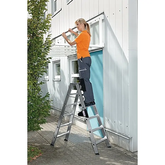 Munk Stehleiter Stehleiter mit ergo-pad beidseitig begehbar 2x3 Stufen - Anzahl Tritte: 3 Stk., Arbeitshöhe: 0,67 m, Belastbarkeit: 150 kg, Aluminium - Silberne Aluminium-Klappleiter, die von einer Frau für Arbeiten in der Höhe an einer Außenwand genutzt wird.