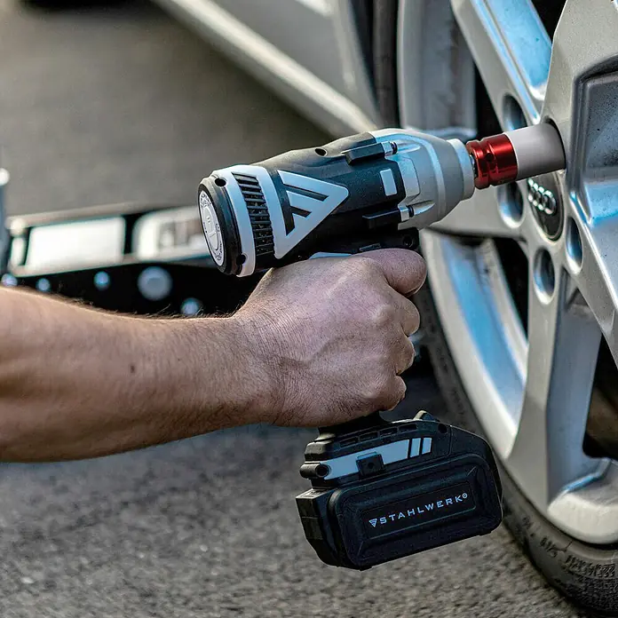 STAHLWERK Akku-Schlagschrauber Drehschlagschrauber ADS-20 ST ohne Akku - 20 V, Ohne Akku, 500 Nm - Eine Hand hält einen schwarz-grauen Akku-Schlagschrauber mit roter Nuss, der eine Radmutter am silbernen Autorad löst.