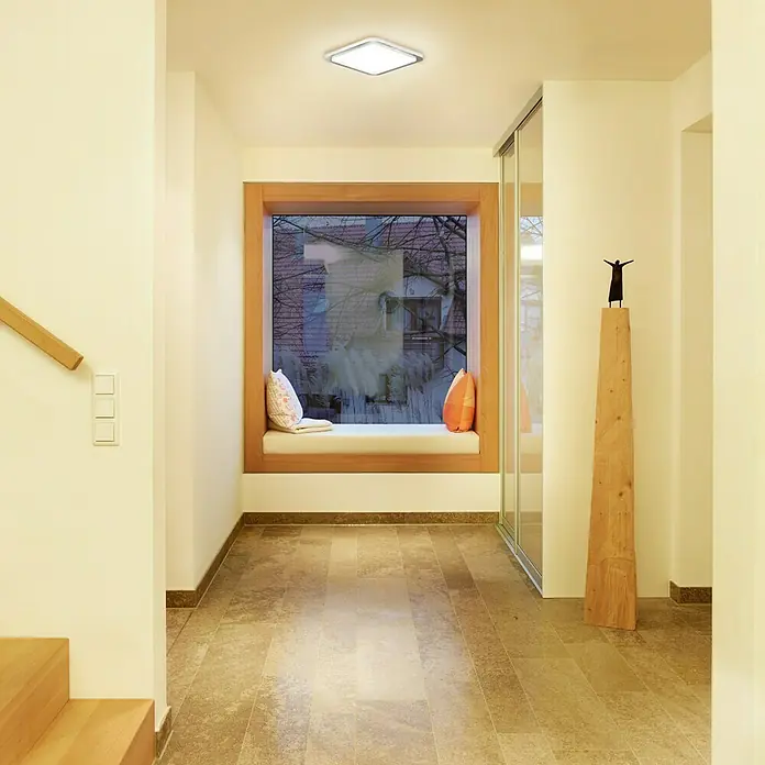 Steinel Sensor-Wand- & Deckenleuchte RS LED D2 S - 8,8 W, L x B x H: 30 x 30 x 6,5 cm, Weiß/Silber - Helle, moderne Fluransicht mit Fliesenboden, Fensterbank mit Kissen und einer quadratischen Deckenleuchte, die den Raum optimal ausleuchtet.