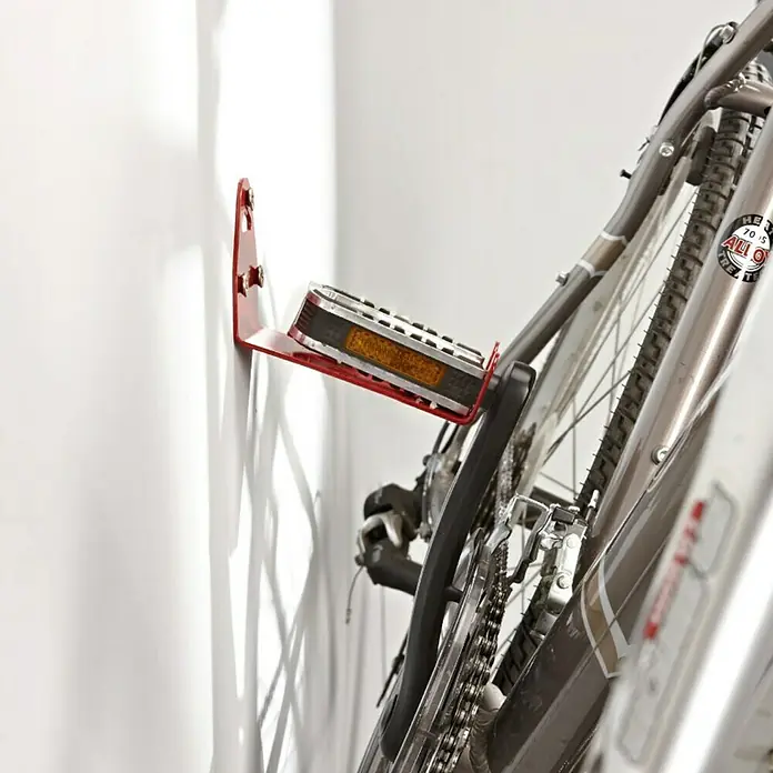 Mottez Gancho de pared para bicicletas B215V - Acero - Un soporte de pared rojo para pedal de bicicleta en una pared blanca, que sostiene una bicicleta. El producto ofrece una solución para ahorrar espacio.