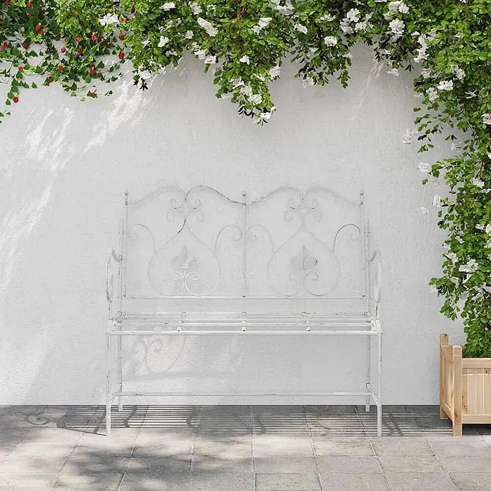 Furnicato Gartenbank Stahl Antik-Weiß - B x T x H: 54 x 104 x 93 cm, Weiß - Weiße, verzierte Metallgartenbank auf gepflasterter Terrasse unter grünen Kletterpflanzen.