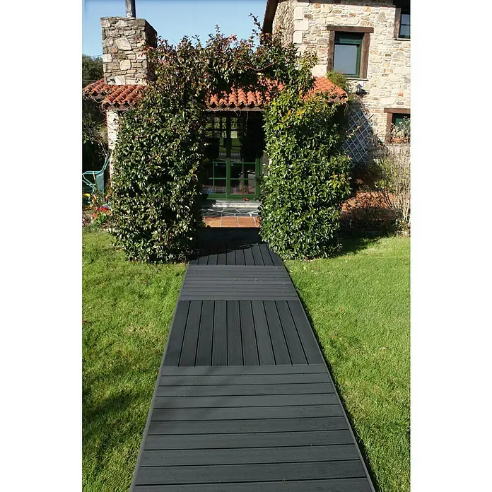 Dioco Loseta de WPC - Antracita, An x L: 90 x 90 cm, 1 ud. - Una pasarela de madera oscura conduce a través de un jardín verde a una casa con una entrada cubierta de vegetación. La pasarela ofrece un paso seguro y elegante.