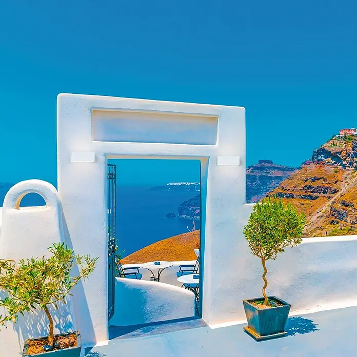 Forlight Aplique exterior Ara - 60 W, 10 x 23 x 9 cm, Blanco, IP65 - Puerta encalada que enmarca una vista azul del mar en Santorini, flanqueada por exuberantes plantas verdes.
