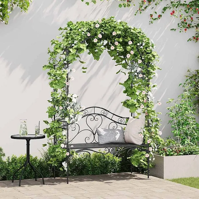 Furnicato Gartenbank Vollstahl - B x T x H: 48 x 121 x 200 cm, Schwarz - Schwarzer Metall-Gartenbogen mit integrierter Bank und künstlichen grünen Ranken, rosa und weißen Blüten, auf einer Terrasse.