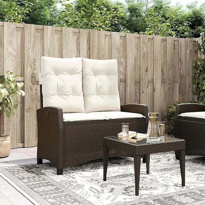 Furnicato Gartenbank - B x T x H: 90 x 90 x 93 cm, Braun und Creme - Braunes Rattan-Gartenmöbel-Set mit weißen Kissen, bestehend aus Zweisitzer-Sofa und Couchtisch, auf einem gemusterten Teppich im Freien.