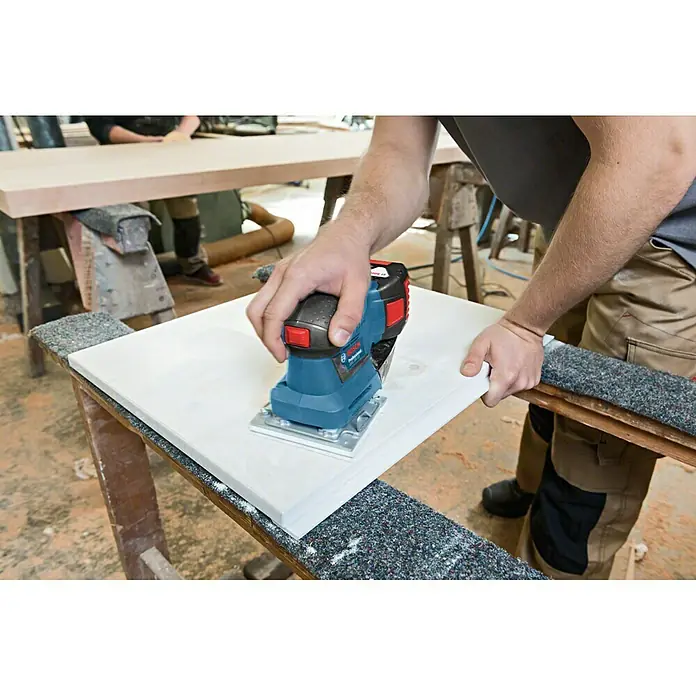 Bosch Professional AMPShare 18V Akku-Schwingschleifer GSS 18V-10 L-Boxx - 18 V, Ohne Akku, Leerlaufschwingzahl: 11.000 U/min - 22.000 U/min - Ein Handwerker benutzt einen Bosch Professional Akku-Multischleifer, um ein Holzbrett zu schleifen. Das blaue Werkzeug ist auf einer Werkbank platziert.
