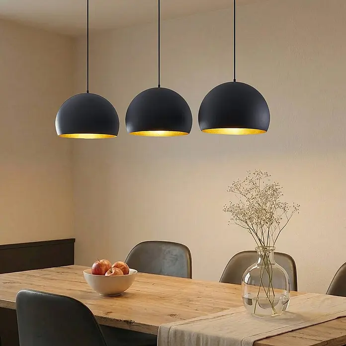 Lindby Pendelleuchte Tarjei - L x B x H: 146 x 40 x 28 cm, schwarz, Schwarz, gold, E27 - Drei schwarze Kuppel-Pendelleuchten mit goldener Innenseite über einem Esstisch aus Holz.