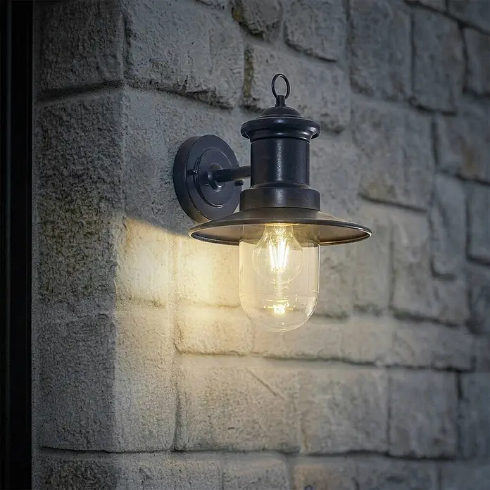 Lindby Außenwandleuchte Eloretta - schwarz, schwarz, IP44 - Dunkle Metall-Außenwandleuchte mit klarem Glasschirm und sichtbarer Edison-LED-Lampe, die eine rustikale Steinmauer beleuchtet.