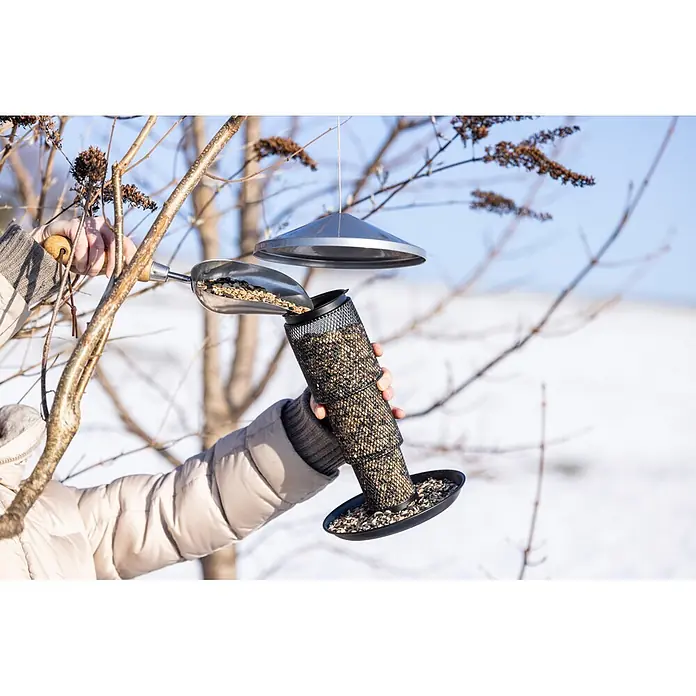 Vogelhaus Rise - Metall, Silber - Schwarzer Gitter-Vogelfutterspender wird mit Vogelfuttermischung aus einer Silberschaufel nachgefüllt.