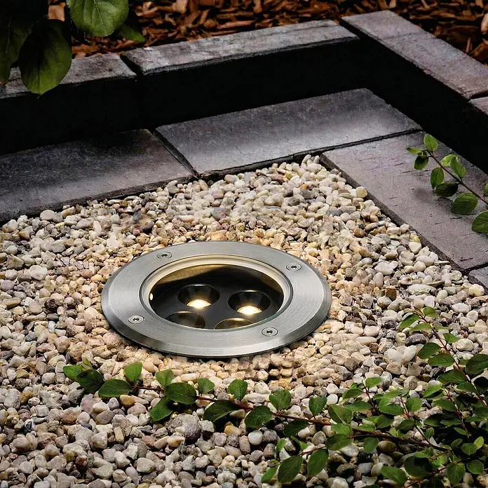 Arcchio LED-Gartenstrahler Thentos - 3.000 K - Einbau-Bodenleuchte aus Edelstahl mit warmer Beleuchtung, in Gartenkies neben Pflastersteinen und Blattwerk.
