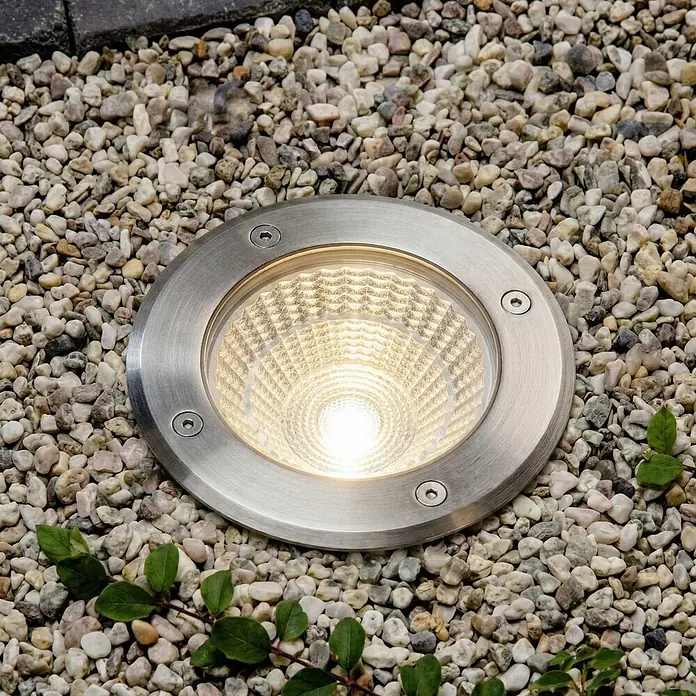 Arcchio LED-Gartenstrahler Thentos - 3.000 K, 12 x 12 x 12,8 cm - Beleuchtete runde silberne Bodeneinbauleuchte mit strukturiertem Reflektor, in hellem Kies eingebettet.
