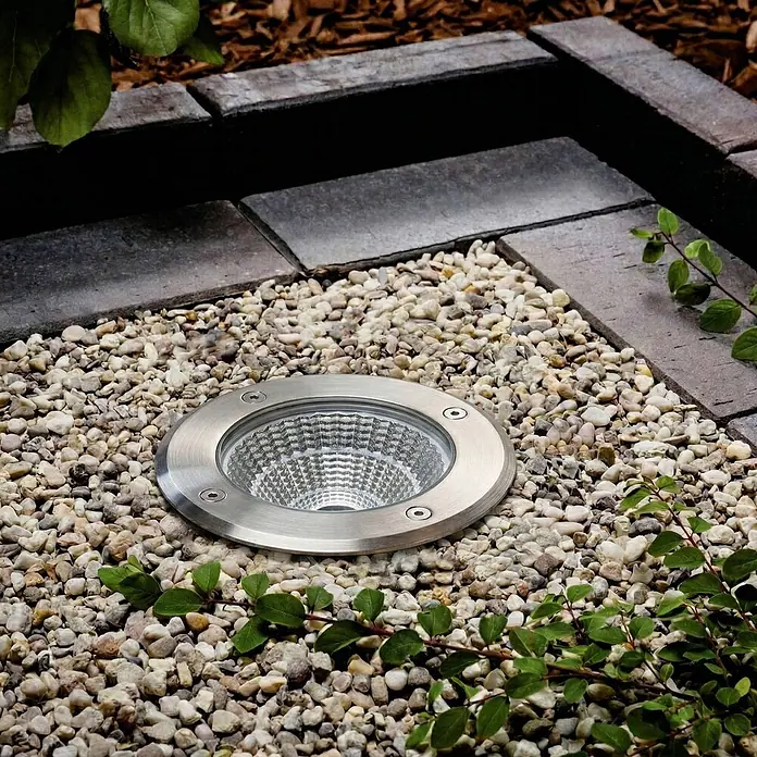 Arcchio LED-Gartenstrahler Thentos - 3.000 K, 12 x 12 x 12,8 cm - Bodenleuchte aus gebürstetem Edelstahl, eingelassen in Kies neben Pflastersteinen und Grünpflanzen.