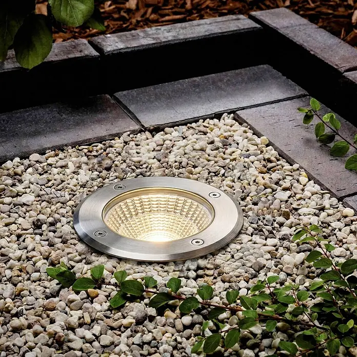 Arcchio LED-Gartenstrahler Thentos - 3.000 K, 12 x 12 x 12,8 cm - Gebürsteter Edelstahl-Bodenstrahler leuchtend in hellem Kies, umgeben von dunklen Pflastersteinen und Grün.