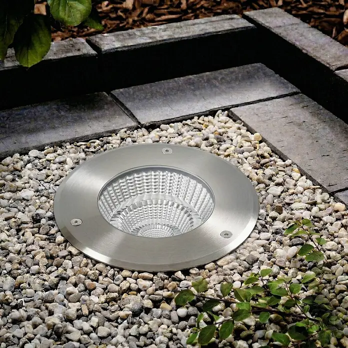 Arcchio LED-Gartenstrahler Thentos - 3.000 K - Rundes Einbau-Außenlicht aus gebürstetem Edelstahl mit texturierter Abdeckung, in Kies neben Pflastersteinen.