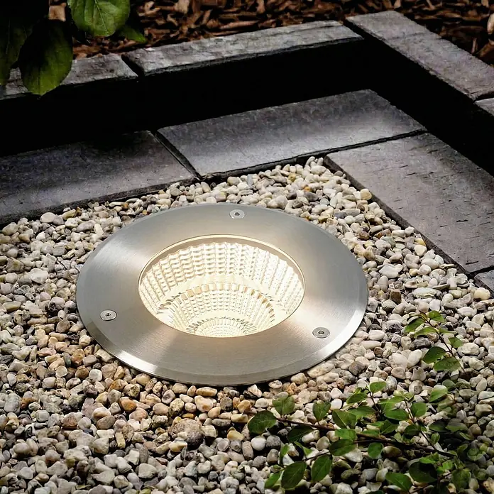 Arcchio LED-Gartenstrahler Thentos - 3.000 K - Runde Einbauleuchte aus gebürstetem Metall, warm leuchtend, in Kies neben Pflastersteinen.