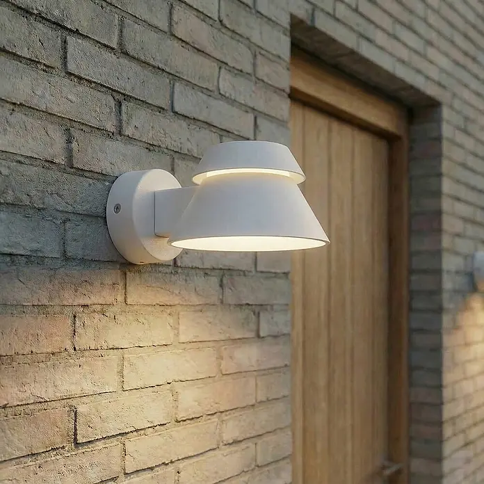 Lucande Außenwandleuchte Moena - 20,7 x 15 x 9,3 cm, alu / grau / zink, alu / grau / zink, IP44 - Weiße Außenwandleuchte an einer Ziegelwand, die warmes Licht abstrahlt.