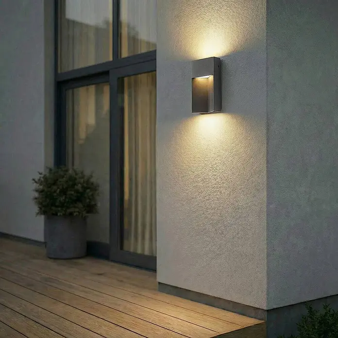Lucande Außenwandleuchte Kaholo - schwarz, schwarz, IP44 - Anthrazitfarbene Außenwandleuchte an einer strukturierten Wand, spendet warmes Auf- und Ablicht auf eine Holzterrasse.