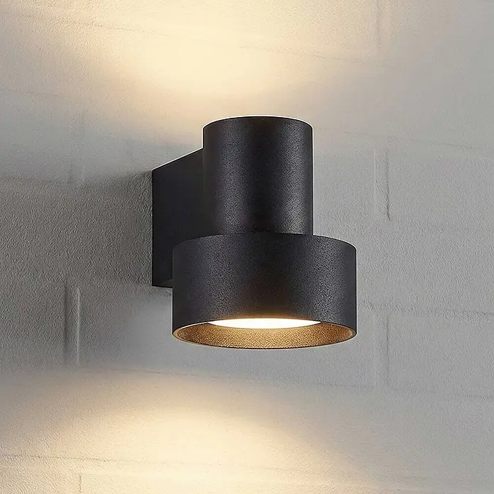Lucande Außenwandleuchte Elune - schwarz, IP44 - Schwarze zylindrische Wandleuchte mit Up/Down-Beleuchtungseffekt auf einer hellen Ziegelwand.