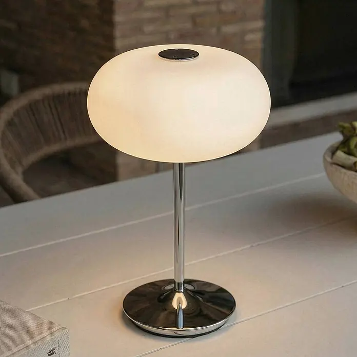 Lucande Tischleuchte Anoria - L x B x H: 18 x 18 x 30 cm, chrom / silber, weiß / opal - Beleuchtete weiße ovale Tischlampe mit Chromständer und -sockel, auf einem hellen Tisch.
